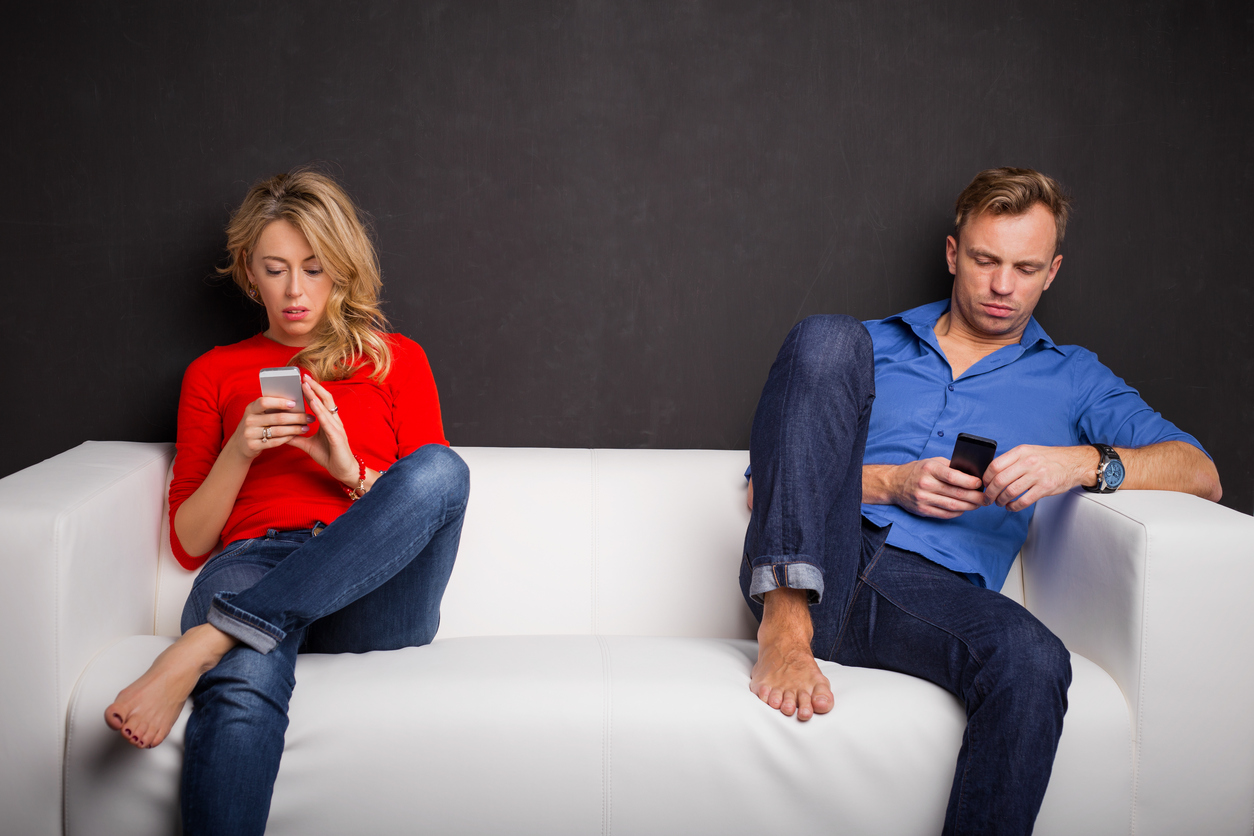 A couple sitting as far as possible from one another on a couch, both on phones