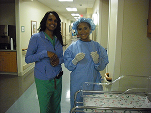 Two students in scrubs and masks pose in front of a nursery