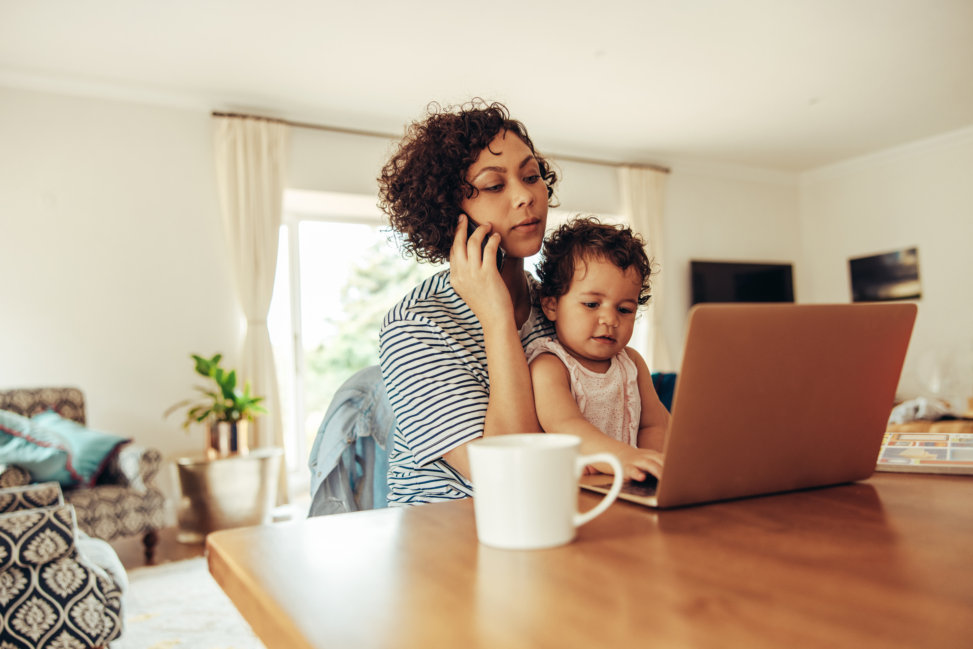 Stock photo of a mother holding a toddler on her lap and sitting in front of the computer while on her cell phone