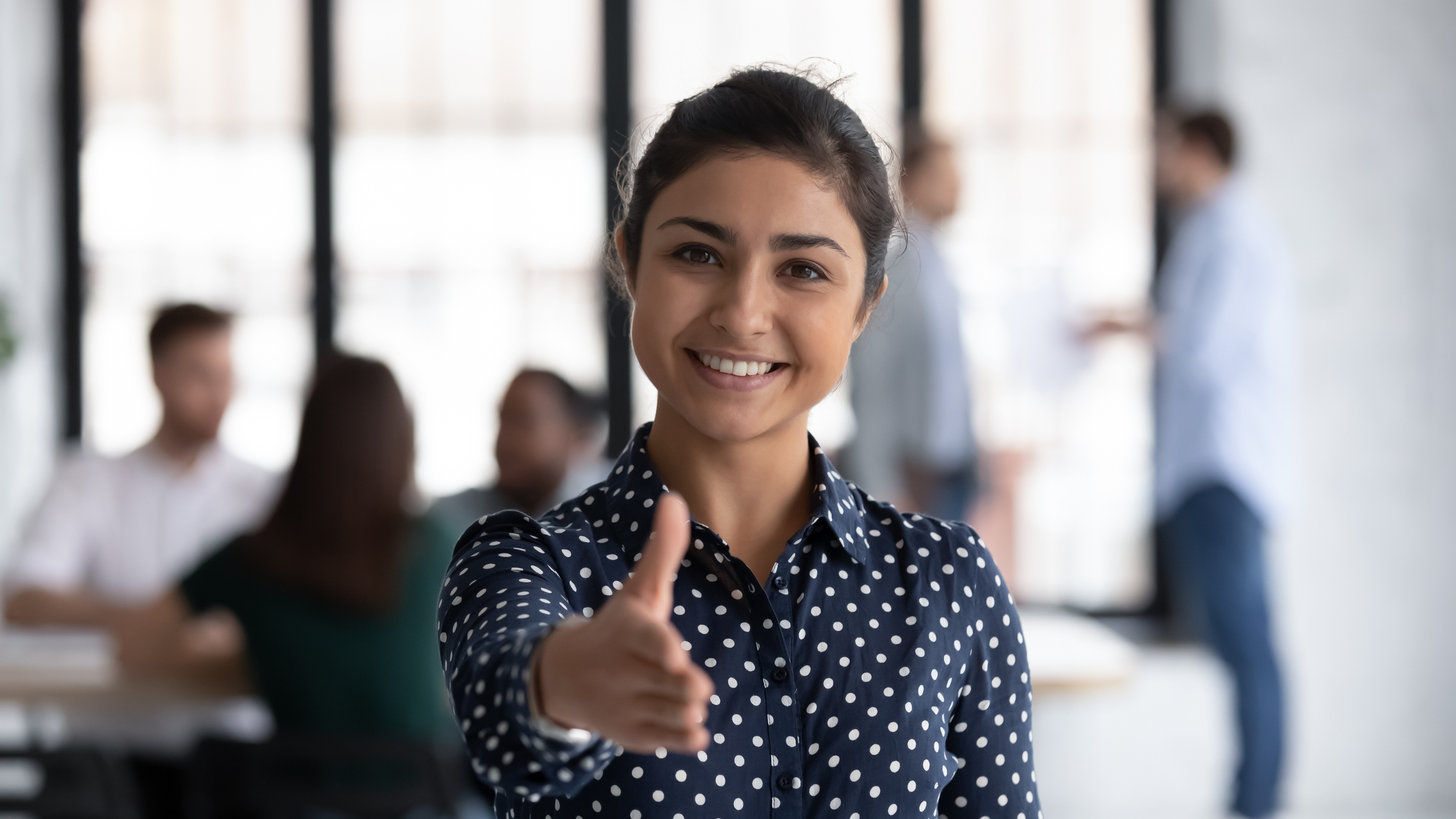 Stock image of an Indian business woman in a busy office extending her hand as to shake hands with the camera person