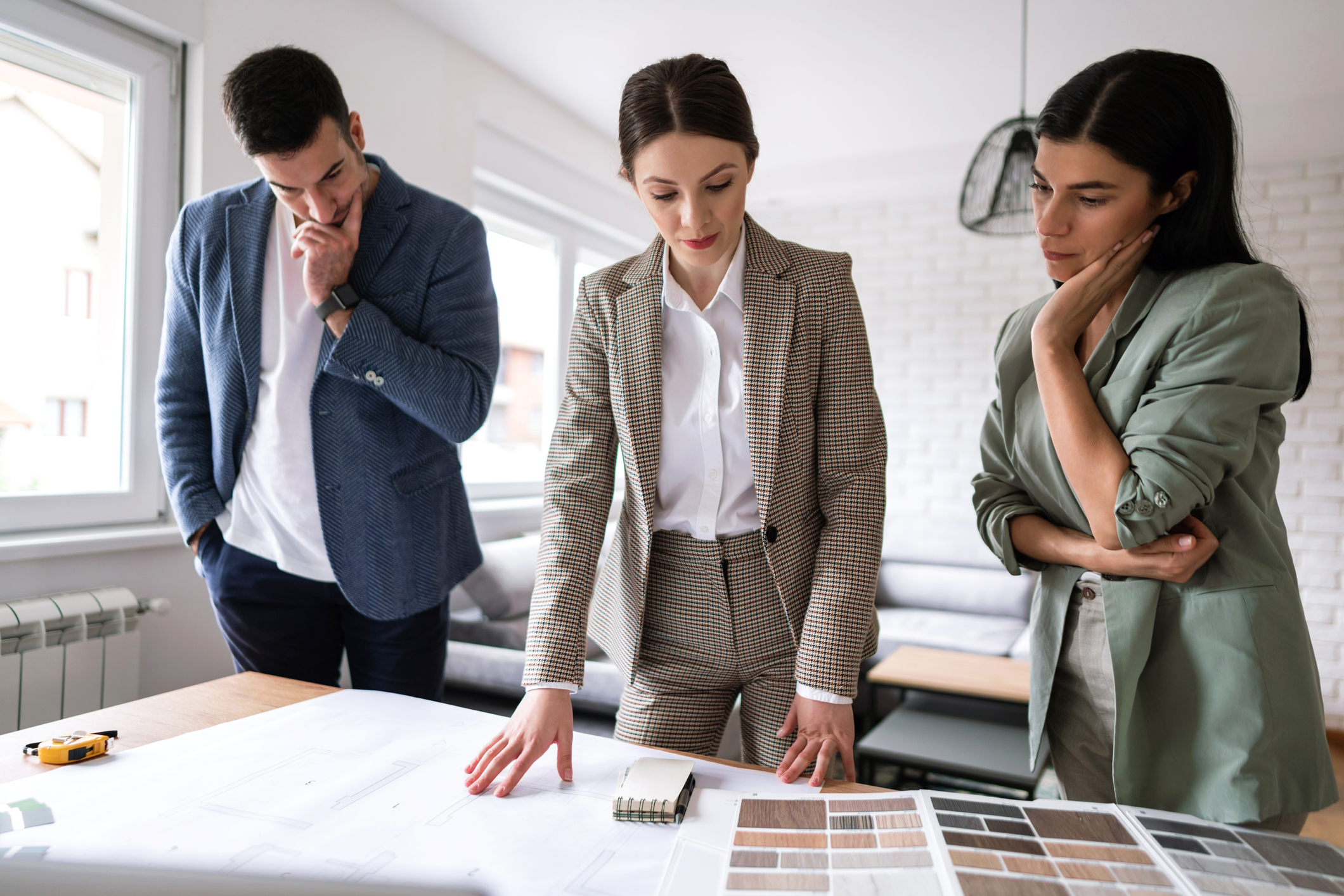 Stock image of a real estate agent standing at a table looking down at house plans and color selections, while a man and woman stand on either side of her with their arms crossed as if in disagreement with the agent