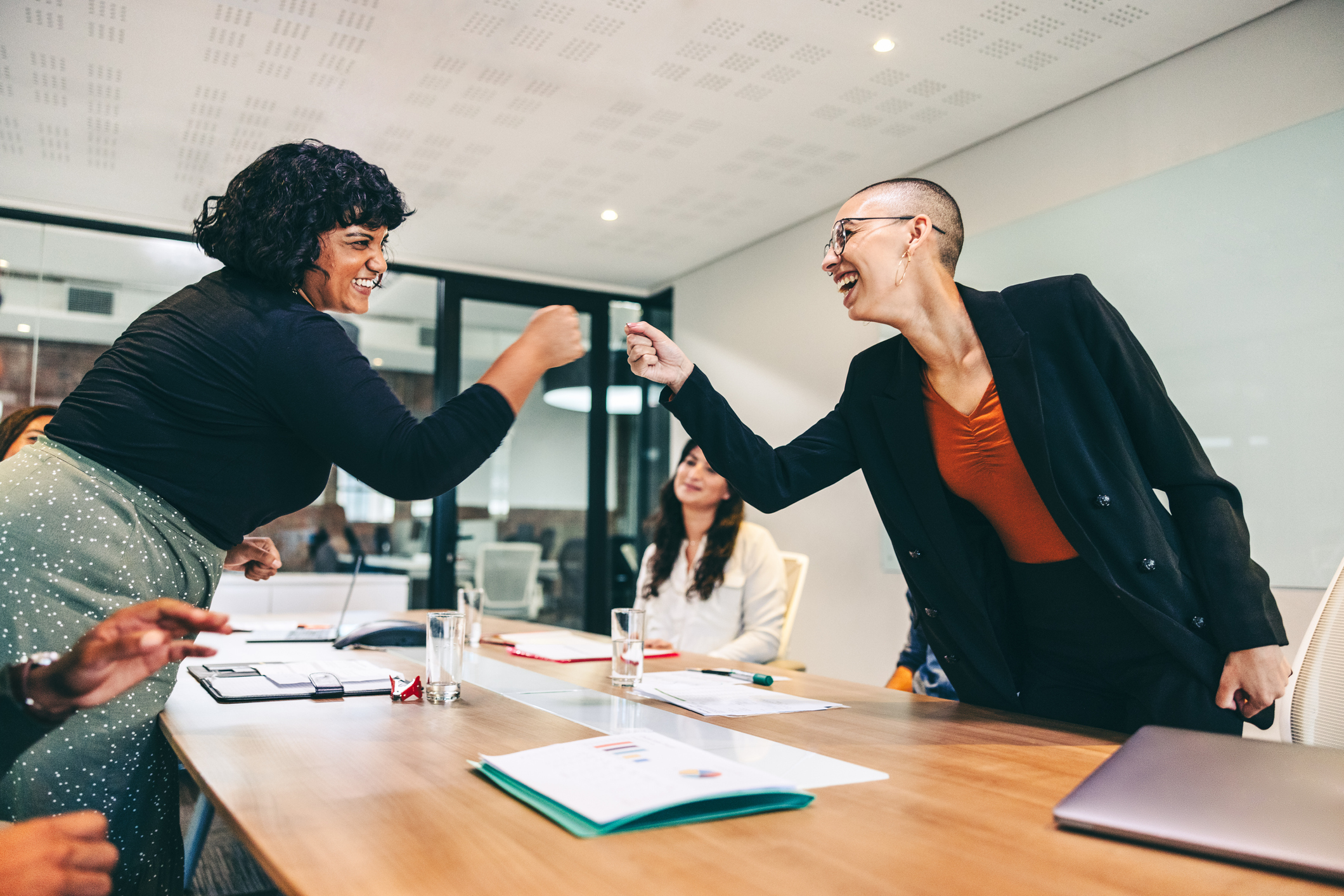 Stock image of two women standing up at conference table and leaning over to give each other a fist bump while smiling