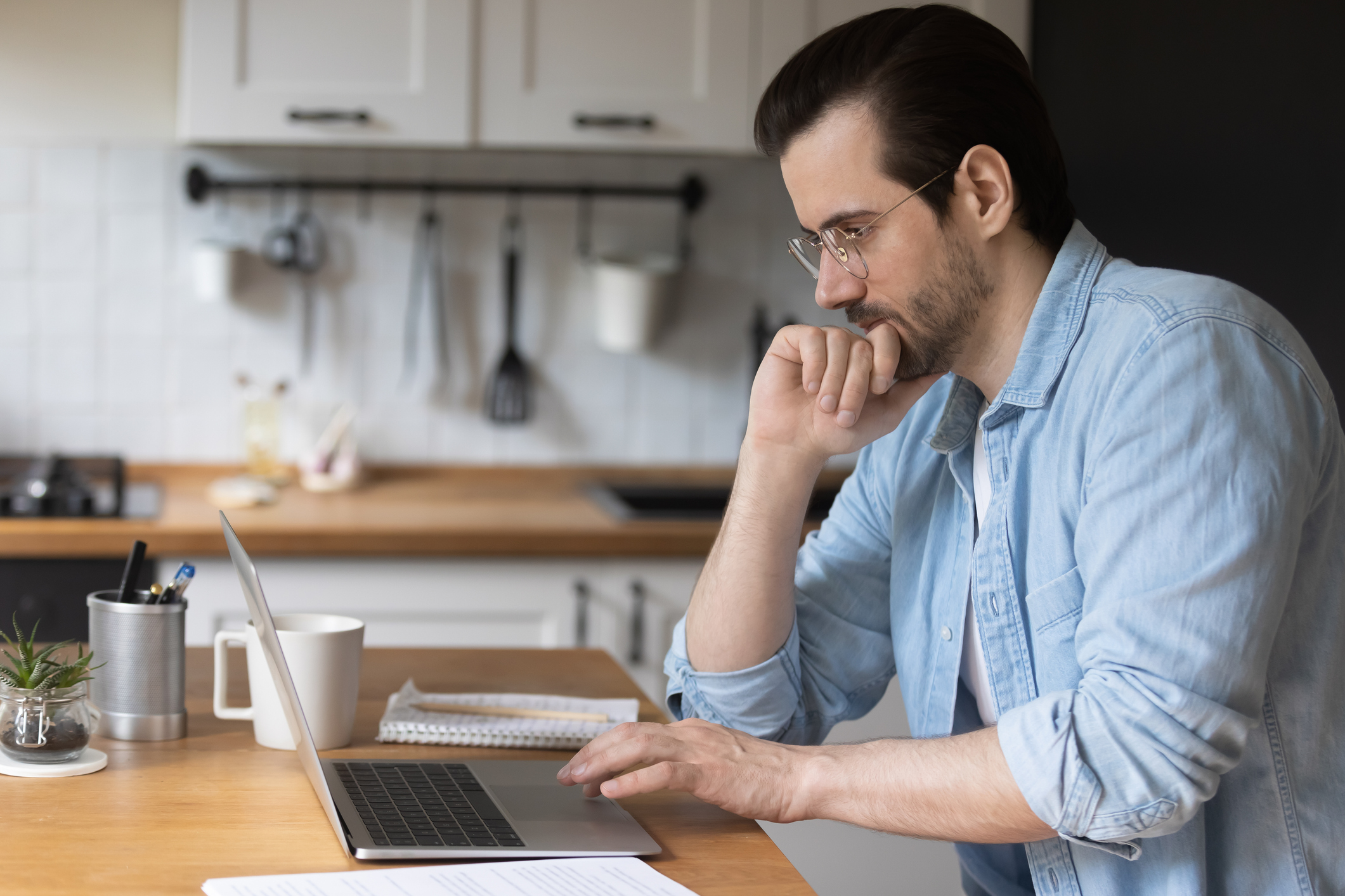 Stock photo of side view of a man sitting in kitchen and looking at his laptop with his right hand under his chin and left hand on the keyboard.