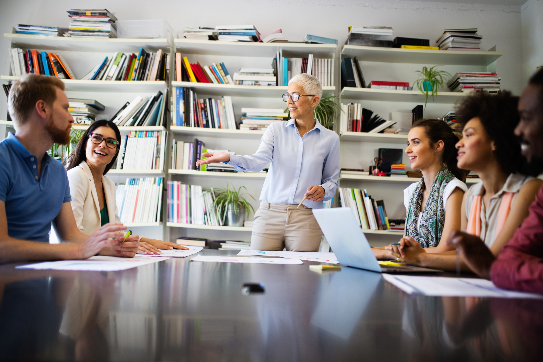 Stock image of team meeting; older woman is standing at the head of the table explaining something to five individuals around the table.