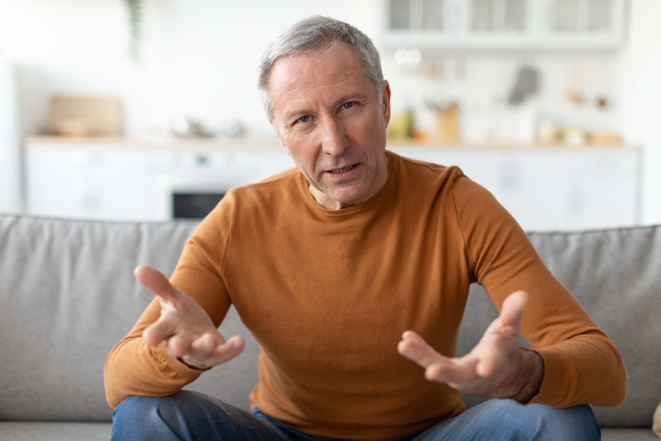 Stock image of older man sitting on couch speaking to camera. His hands are outstretched like he is talking with his hands