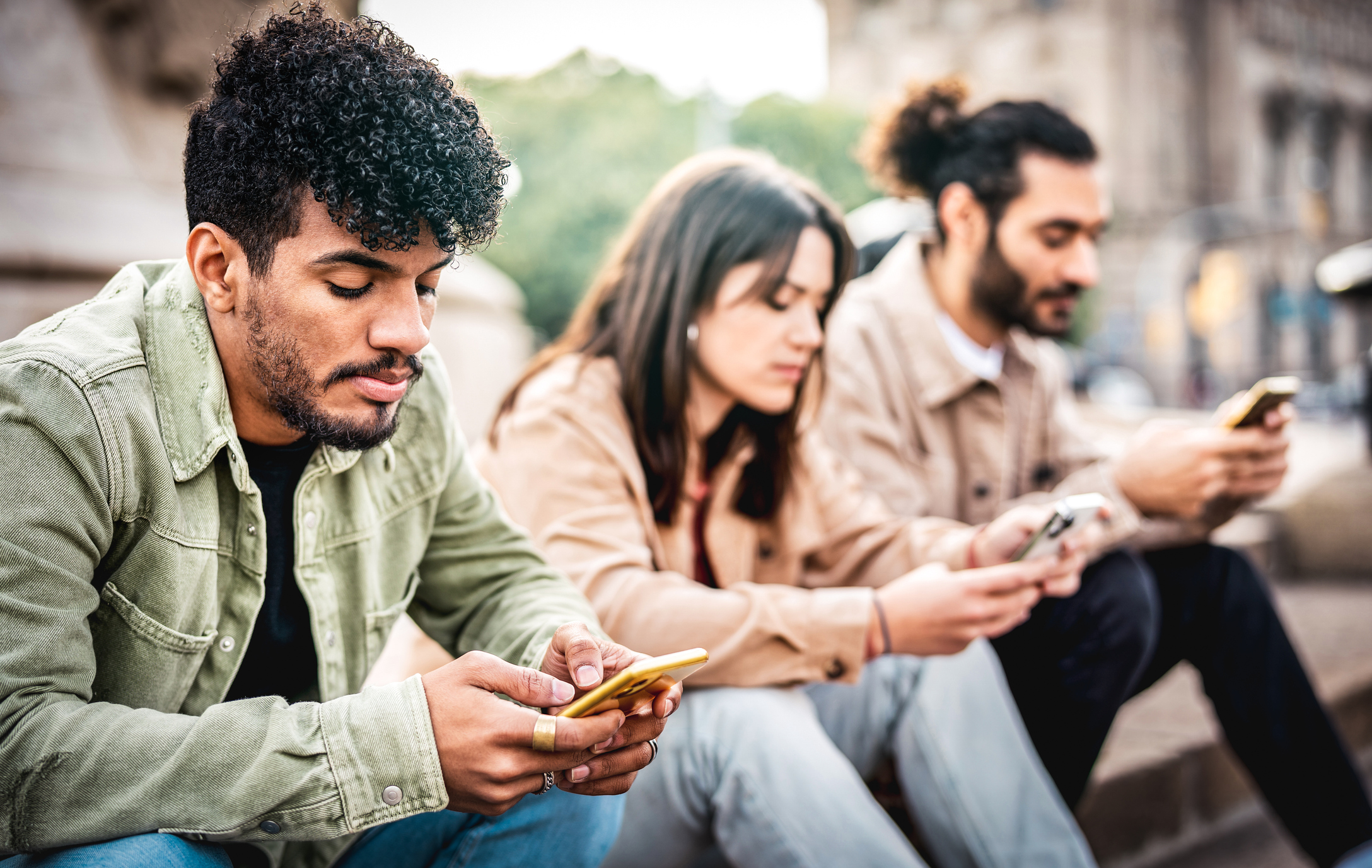 Stock image of three people, man, woman, man, sitting next to one another and all looking at their phones.